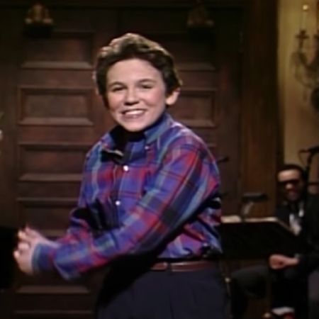 Fred Savage is smiling and talking to the audience as musicians are sitting in the background.